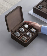 A person in a blue shirt is arranging the TAWBURY Fraser 6 Watch Case - Brown, containing wristwatches with diverse designs and metal straps. Another watch case is visible in the background, providing perfect storage for collectors.